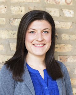 Clare is a white woman with shoulder length dark brown hair. She stands in front of a brick wall, wearing a blue shirt with a gray blazer, and smiles at the camera.