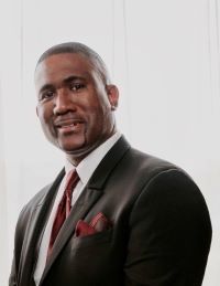 Darryl smiles in front of a white background and is wearing a black suit with a red tie. 