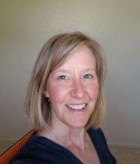 JP blonde shoulder-length hair, grey eyes smiles in front of a white wall and is wearing a dark blue blouse.