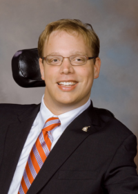 A photo of a 32-year-old white male smiling with glasses and curly brown hair sitting in his wheelchair wearing a black suit, a white shirt, and a red, blue, and white striped tie.